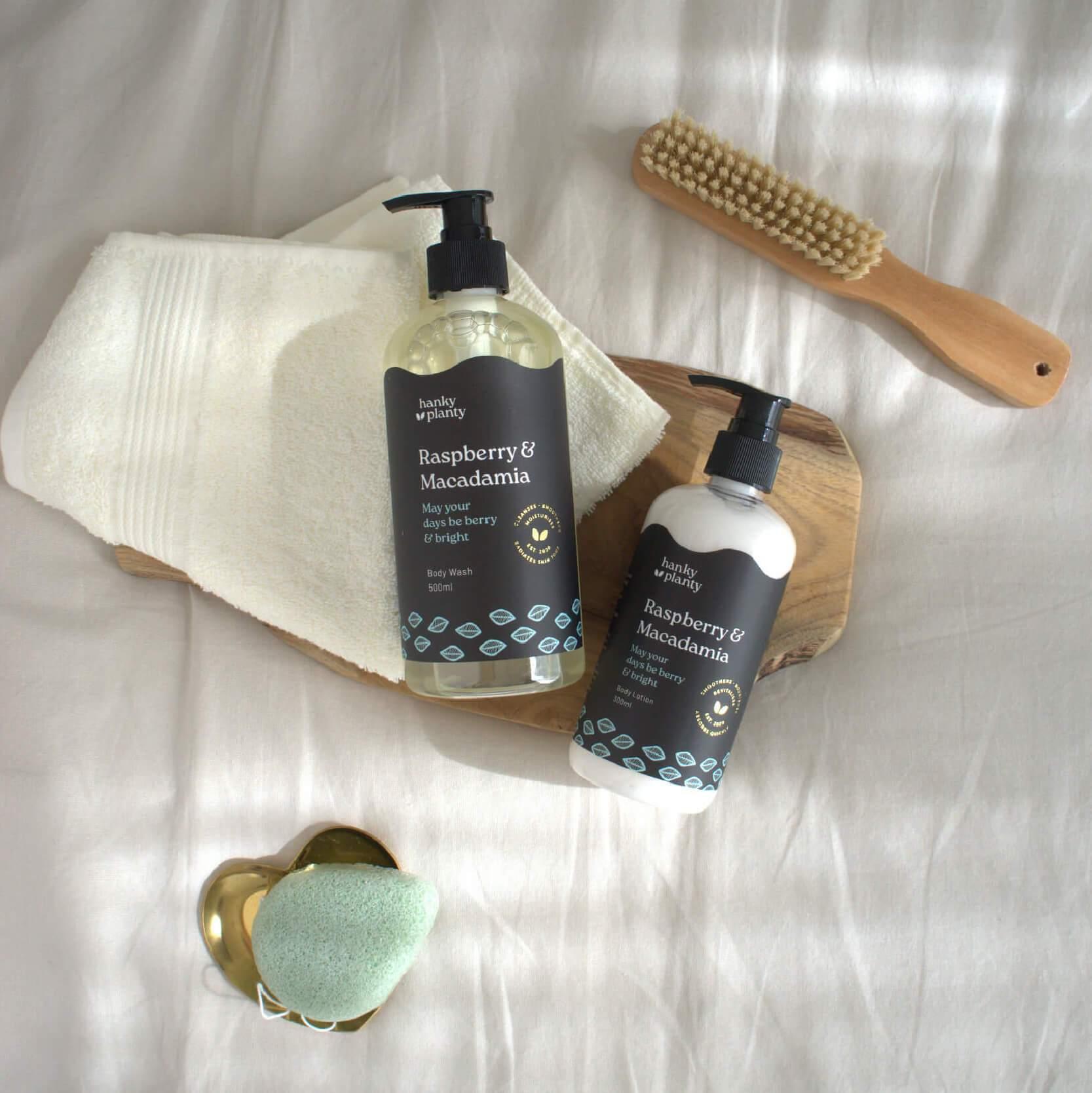 Hanky Planty Raspberry & Macadamia natural body wash and body lotion, 500ml pump bottles with black labels, displayed on a towel and wooden tray with bath brush and konjac sponge.