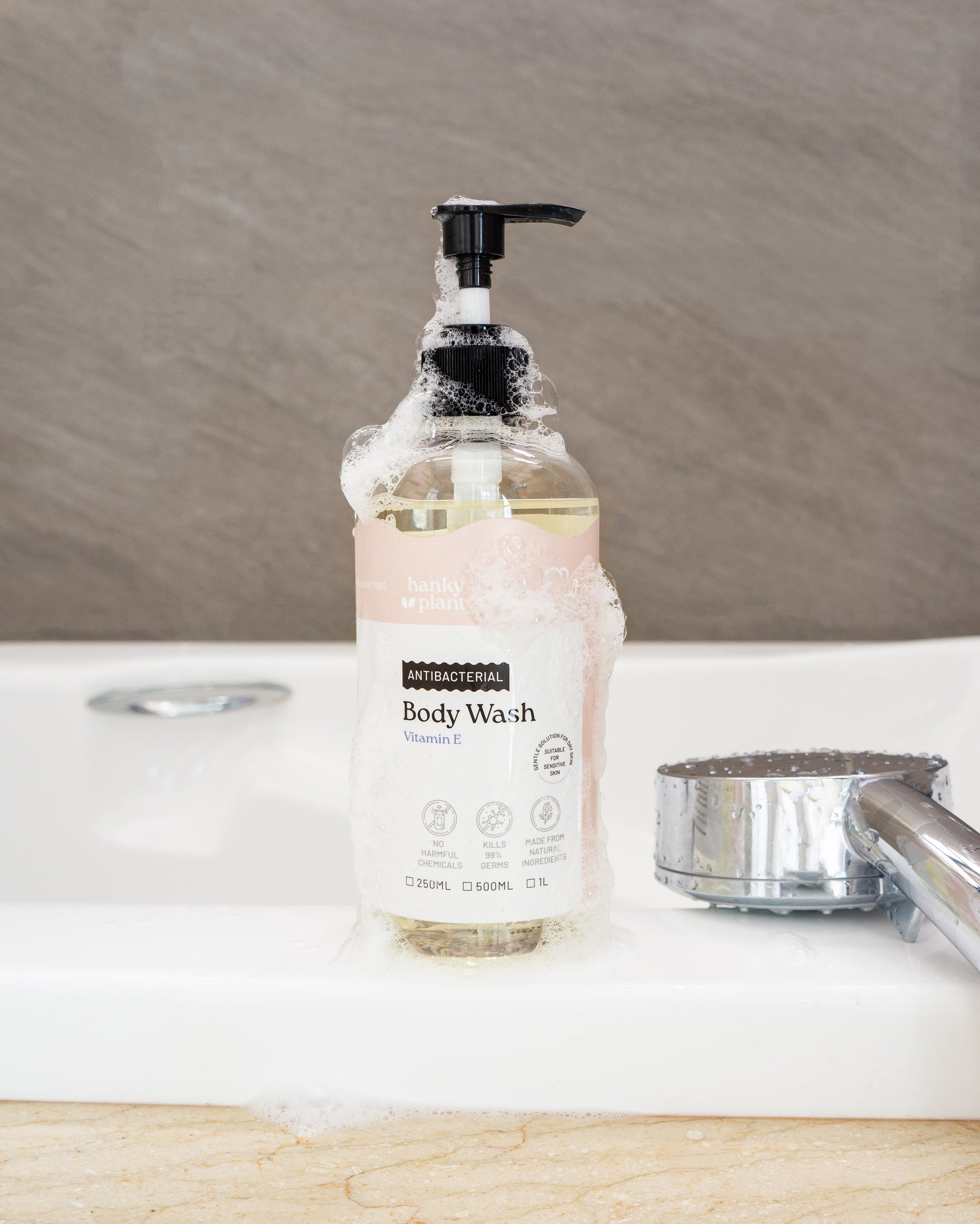 Hanky Planty Antibacterial Body Wash with Vitamin E, 500ml pump bottle, covered in foamy lather, placed on a bathroom sink beside a shower head.