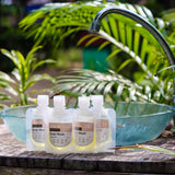 Hanky Planty Petite Antibacterial Body Wash collection in 30ml refill pouches, assorted scents, displayed outdoors on a wooden surface beside a green basin with tropical plants in the background.