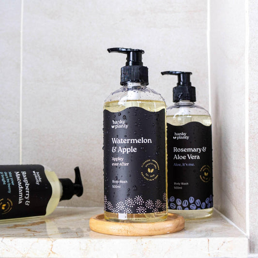 Hanky Planty natural body wash collection, featuring 500ml Watermelon & Apple, Rosemary & Aloe Vera, and Raspberry & Macadamia variants in black-labeled pump bottles with golden liquid, displayed in a tiled bathroom.