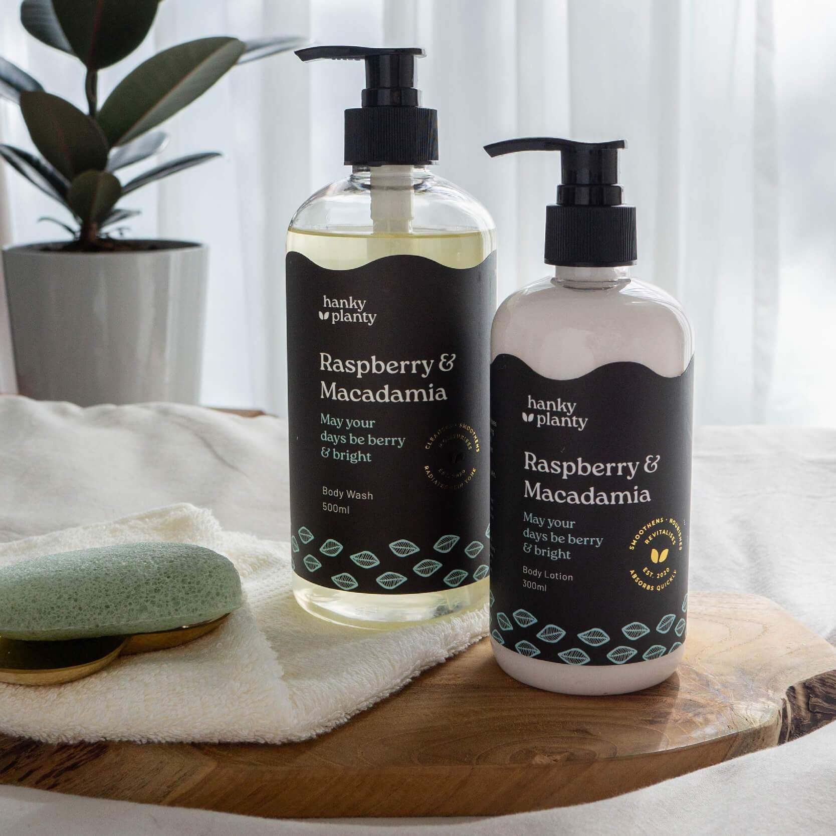 Hanky Planty Raspberry & Macadamia set, featuring 500ml natural body wash and 300ml body lotion in black-labeled pump bottles, displayed on a wooden tray with a towel and green konjac sponge by a window.