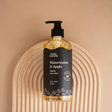 Hanky Planty Watermelon & Apple natural body wash, 500ml pump bottle with black label and golden liquid, placed on an arched wooden board against a beige background.
