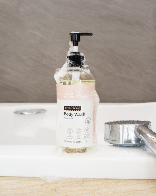 Hanky Planty Antibacterial Body Wash with Vitamin E, 500ml pump bottle, covered in foamy lather, placed on a bathroom sink beside a shower head.